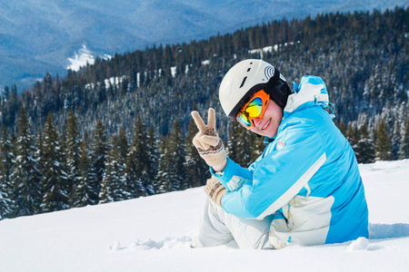 穿着蓝色西装头盔和眼镜的妇女坐在山顶上滑雪板附近的雪地上。