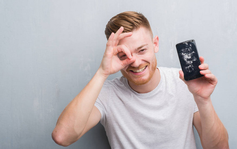 灰色垃圾墙上的年轻红头发的人持有破碎的智能手机与快乐的脸微笑做 ok 标志用手在眼睛看通过手指