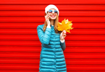 时尚秋季微笑的女人在智能手机上举行的谈话在城市的红色背景手捧着黄枫树叶