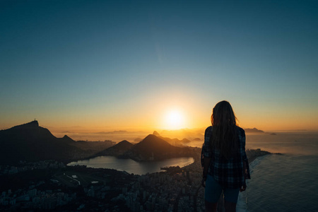 巴西, 里约热内卢。在山上遇见黎明两兄弟。看大城市, 湖, 海, 山, 山。橙色蓝色的颜色剪影的女孩谁看着城市和太阳