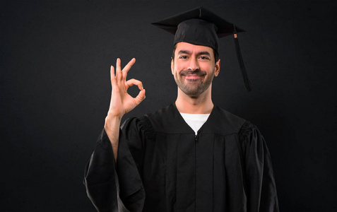 在毕业日大学的男子用手指显示一个 ok 标志。在黑色背景下面对幸福和满足