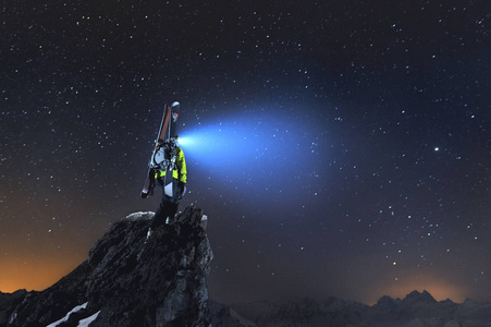 夜风景。一个专业的野外滑雪者, 背包和滑雪板矗立在山上的岩石上, 并在天空中闪耀着车头灯。