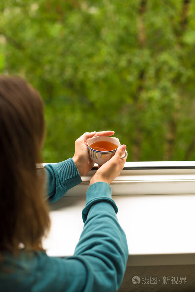 平静的黑发年轻女孩静静地看着打开的窗户, 手里拿着一杯茶