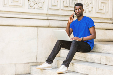 年轻的非洲裔美国大学生在纽约学习, 穿着蓝色 t恤衫, 黑色裤子, 运动鞋, 手表, 坐在校园的楼梯上, 在笔记本电脑上工作, 