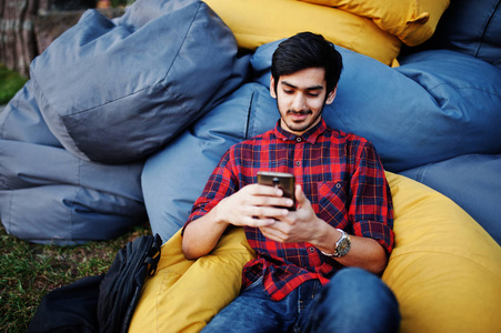 年轻的印度学生男子在格子衬衫和牛仔裤坐在户外枕头放松。用手机消磨时间
