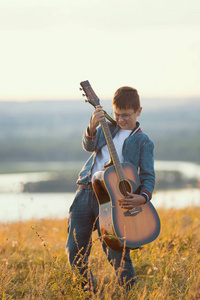 年轻的微笑的男孩演奏吉他在夏天领域在日落, 远摄射击图片