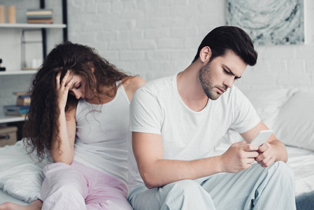 年轻男子使用智能手机, 而心烦的女人坐在床后, 关系困难的概念