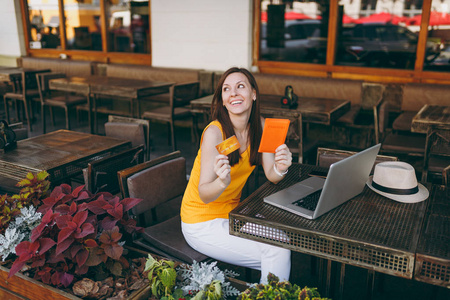妇女在户外街咖啡厅坐在餐桌上, 用现代笔记本电脑计算机手持银行信用卡护照订票机票。餐馆在空闲时间。生活方式经营理念