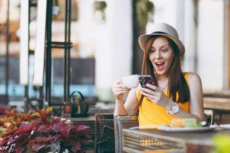妇女在户外街道咖啡馆咖啡馆坐在帽子与杯子卡布奇诺蛋糕, 使用手机, 放松在餐馆在空闲时间。移动办公室在夏天。生活方式休息概念