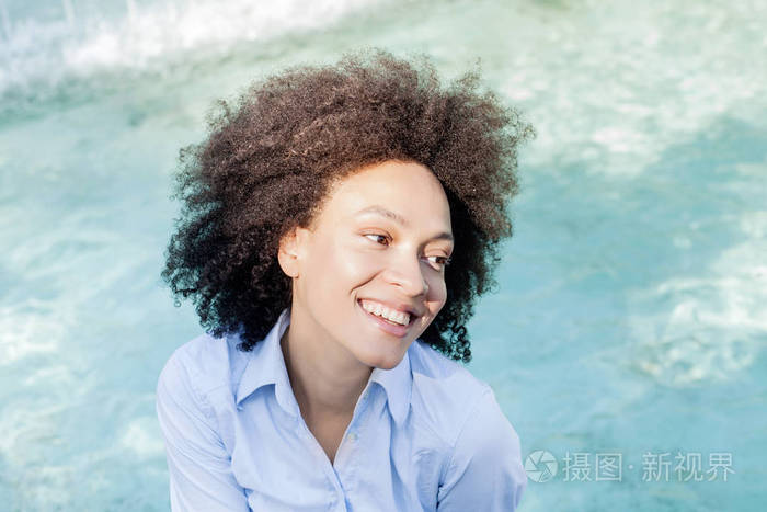 美丽快乐的美国黑人年轻女子肖像, 微笑混合的种族女性放松附近的喷泉, 看着侧面, 阳光反射在脸上