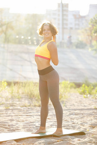 运动的年轻女子伸展户外瑜伽垫穿着灰色健身房裤和黄色运动顶部穿。美丽的白色女孩与肌肉做前锻炼伸展前锻炼