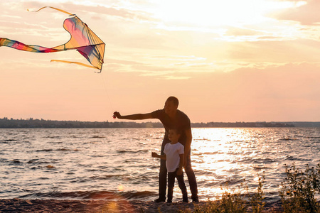 快乐的父亲和儿子在日落时在河边放飞风筝