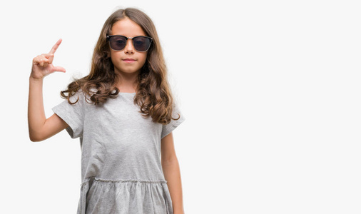 黑发西班牙裔女孩戴着太阳镜微笑着, 自信的手势与手做尺寸标志, 而在看和相机。度量概念