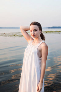 黎明时分站在河边的白色礼服的迷人女孩。美丽, 时尚, 肖像