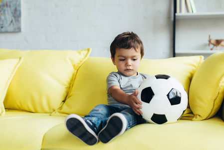 可爱的男孩坐在黄色沙发与足球
