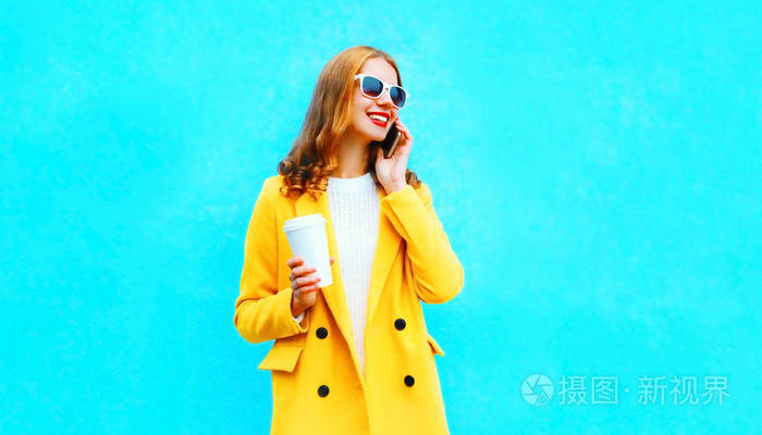 时尚肖像微笑的女人谈智能手机持有彩色蓝色背景杯咖啡