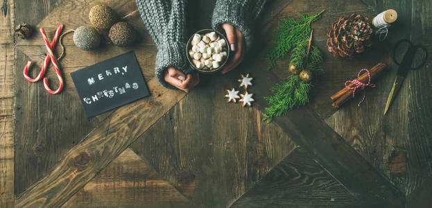 圣诞, 新年准备背景。平躺的贺卡, 闪闪发光的玩具, 女人的手在毛衣拿着一杯热巧克力, 肉桂, 剪刀在木桌上