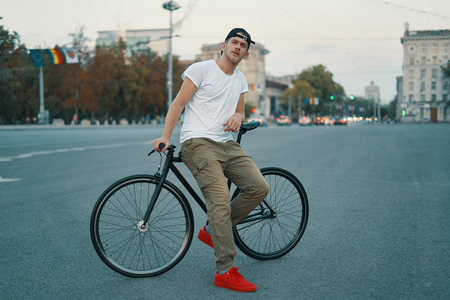 在街上的一个现代年轻人的户外肖像, 在自行车上休息。一个年轻的运动男子身穿卡其裤, 白色 t恤, 红色运动鞋和背包探索欧洲城市