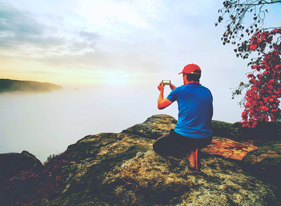 坐在岩石顶峰的勇敢的人在他的手机上拍照。秋天自然