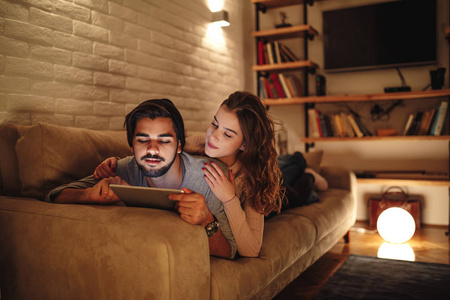 一对年轻夫妇躺在沙发上, 因为男友正在浏览一块平板电脑看电影。