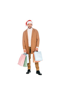 英俊的胡子年轻男子在圣诞老人帽子举行购物袋, 微笑着在白色的相机隔离