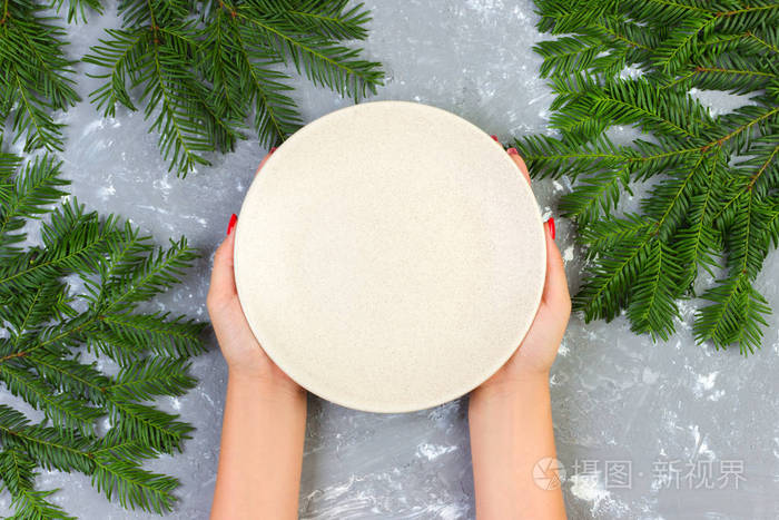女性手拿着一个空白色盘子在灰色背景与圣诞节分支.