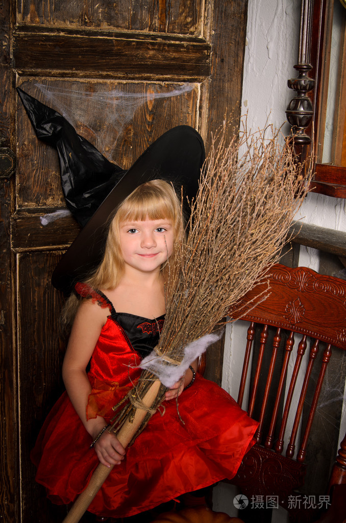 可爱的金发女郎万圣节女巫红色礼服和黑色帽子在旧木门室内扫帚
