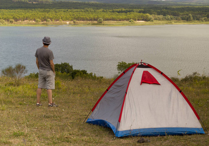 旅行者的帽子是站在帐篷附近, 看着美丽的湖景在一个夏天的一天。野营生活方式户外度假概念