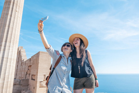 两个年轻美丽快乐的女人女性旅行女友在希腊的海洋和古遗址的背景下做自拍