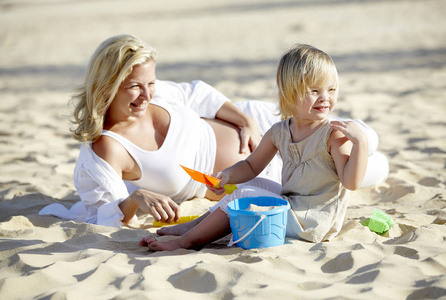 年轻的孕妇和女儿坐在沙滩上玩沙滩玩具