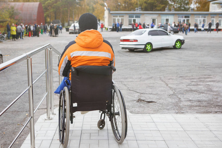 残疾人在轮椅上的橙色背心坐在门廊上, 他的背部对观众和看着汽车