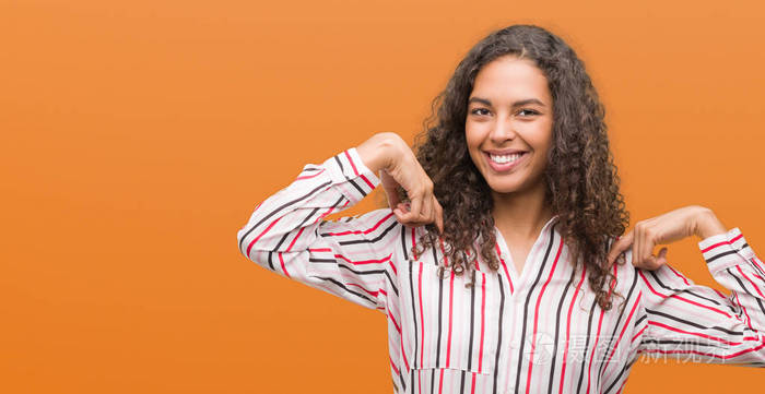 美丽的年轻西班牙裔妇女看着自信与微笑在脸上, 指着自己的手指自豪和