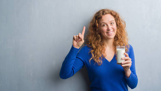 年轻的红头发的妇女喝玻璃牛奶惊讶的想法或问题指向手指与快乐的脸, 头号