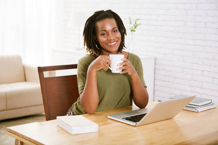 愉快的妇女坐与杯子咖啡在工作桌与膝上型电脑和日志在家里