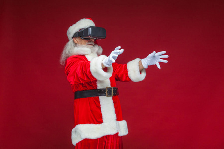 圣诞节。圣诞老人在黑色虚拟现实眼镜做手势用他的手。惊喜情绪新技术。在红色背景上被隔离