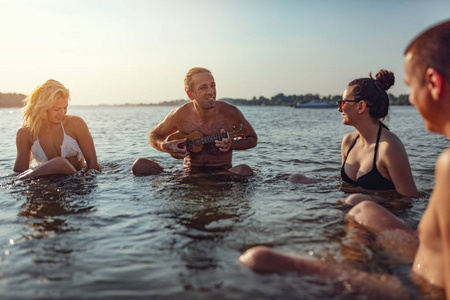 一群快乐的朋友坐在水和人玩四弦琴在日落
