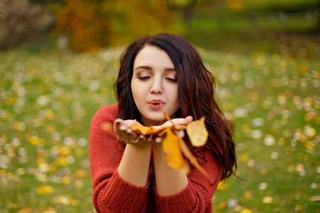 红色针织 sweather 的年轻美女 Portait 躺在草地上, 从她的手在秋季公园的叶子上吹树叶黄色的叶子