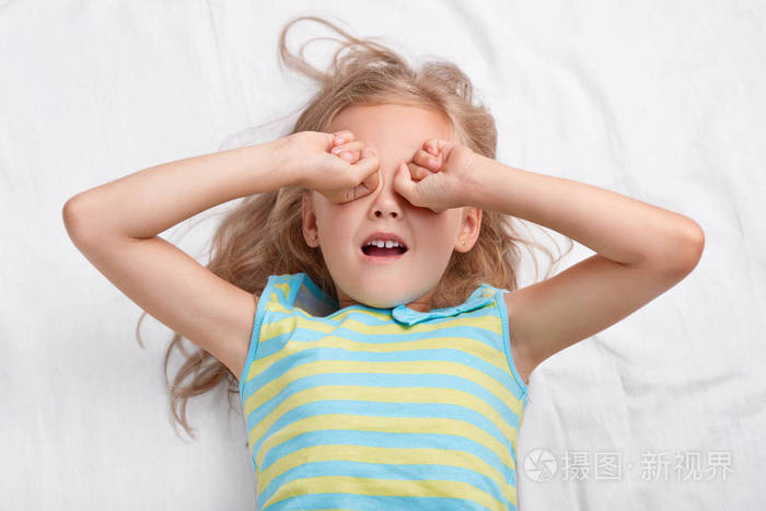 照片中的漂亮的浅色头发的女孩揉眼睛, 想睡觉, 穿着睡衣, 躺在舒适的