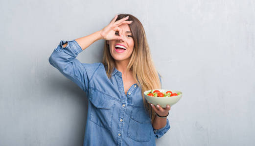 灰色垃圾墙上的年轻成年妇女吃健康的西红柿沙拉与愉快的面孔微笑做 ok 标志用手在眼睛看通过手指