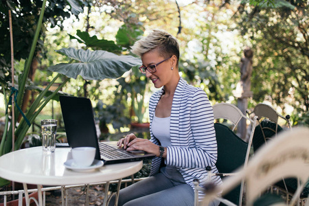 年轻快乐的女人坐在美丽的花园像咖啡馆酒吧或餐厅, 并在她的笔记本电脑上做一些事情