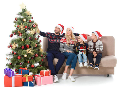 快乐的家庭在圣诞老人的帽子坐在沙发附近的圣诞树与礼物, 孤立的白色