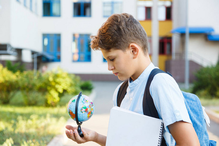 可爱的小男孩穿着蓝色衬衫, 背包和工作簿在他的学校前面握着地球仪。教育, 回到学校概念