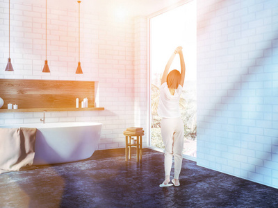 穿着睡衣的妇女站在白色砖浴室角落里, 有一个黑色浴缸和一个热带景观的全景窗口。色调图像