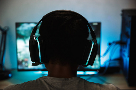 后视图特写年轻的玩家男孩玩电子游戏在线电脑在黑暗的房间里戴着耳机的麦克风