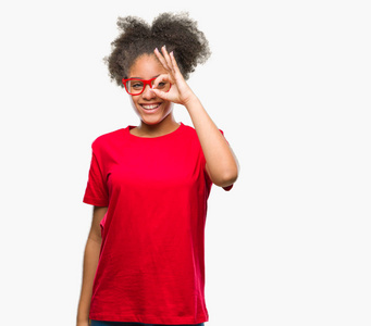 年轻的美国黑人妇女戴眼镜在孤立的背景下做 ok 手势用手微笑, 眼睛看通过手指与愉快的面孔