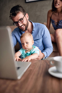 微笑繁忙的父亲工作与计算机, 当照看他的婴孩儿子时