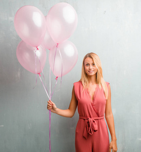 优雅的年轻金发女子持有粉红色气球对墙壁