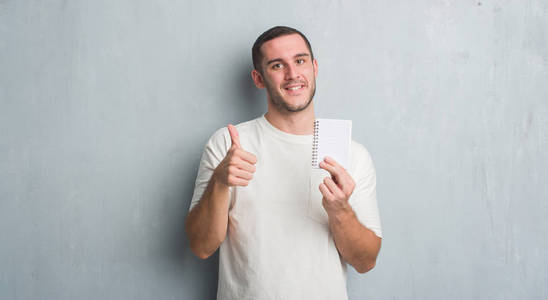 年轻的白种人在灰色垃圾墙显示笔记本愉快的大微笑做 ok 标志, 拇指与手指, 优秀的标志