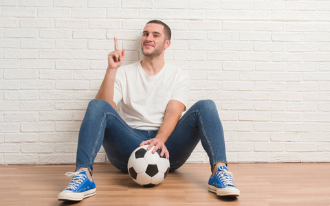 年轻的白种人坐在白色砖墙持有足球球惊讶于一个想法或问题指向手指与快乐的脸, 第一