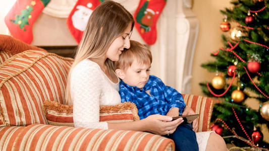 小蹒跚学步的男孩坐在圣诞树旁边, 看着手机上的卡通片